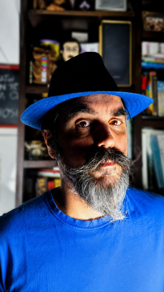 Stefano Giolo in primo piano con cappello nero a tesa blu, barba lunga e maglietta blu, davanti a una libreria